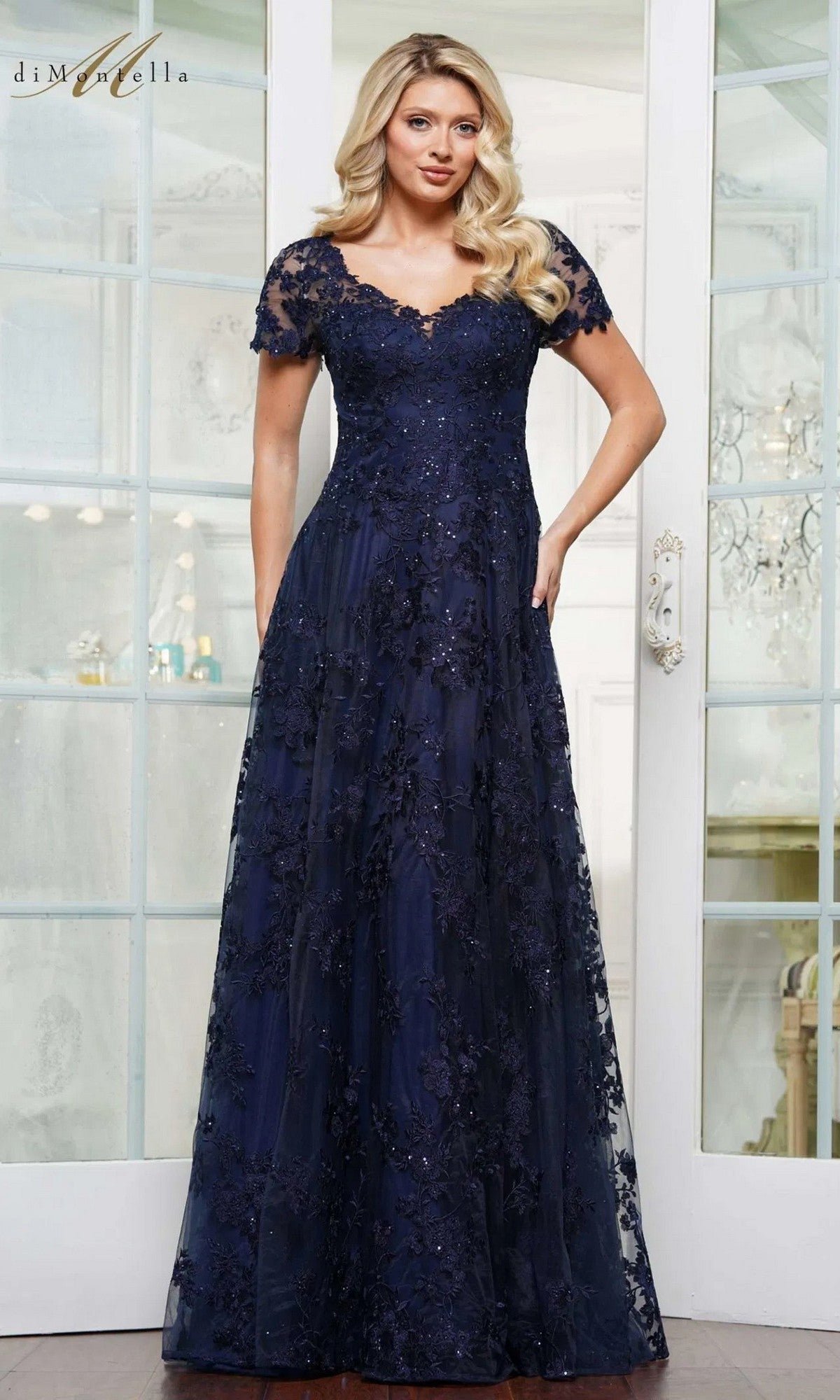 A woman stands indoors wearing the Rina Di Montella RD3248 long formal dress in navy, featuring short sleeves, a floor-length a-line skirt, beaded floral embroidery, and sequin accents.